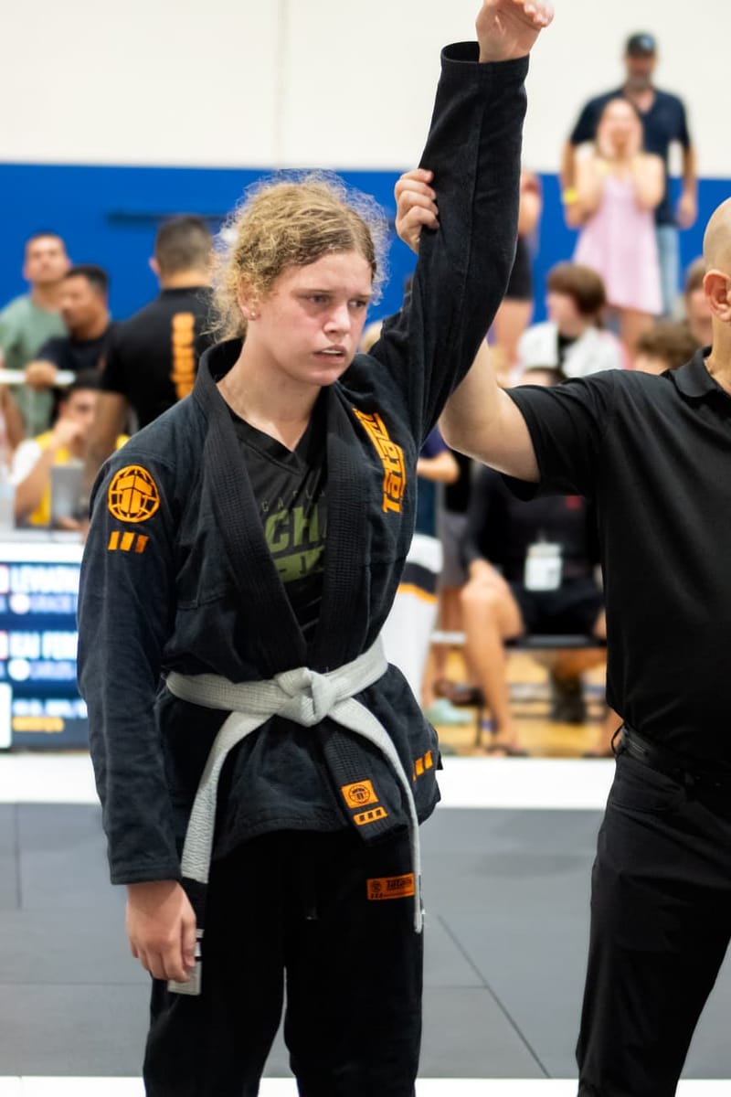 A teenage girl in a black gi has her arm raised by a referee after winning a jiu-jitsu match.