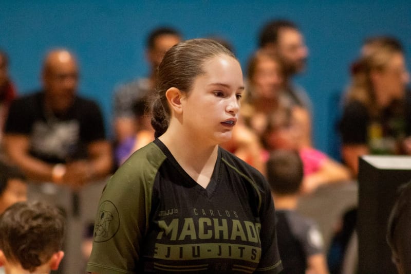 A teen girl in a Machado rashguard waits matside with the crowd behind her.
