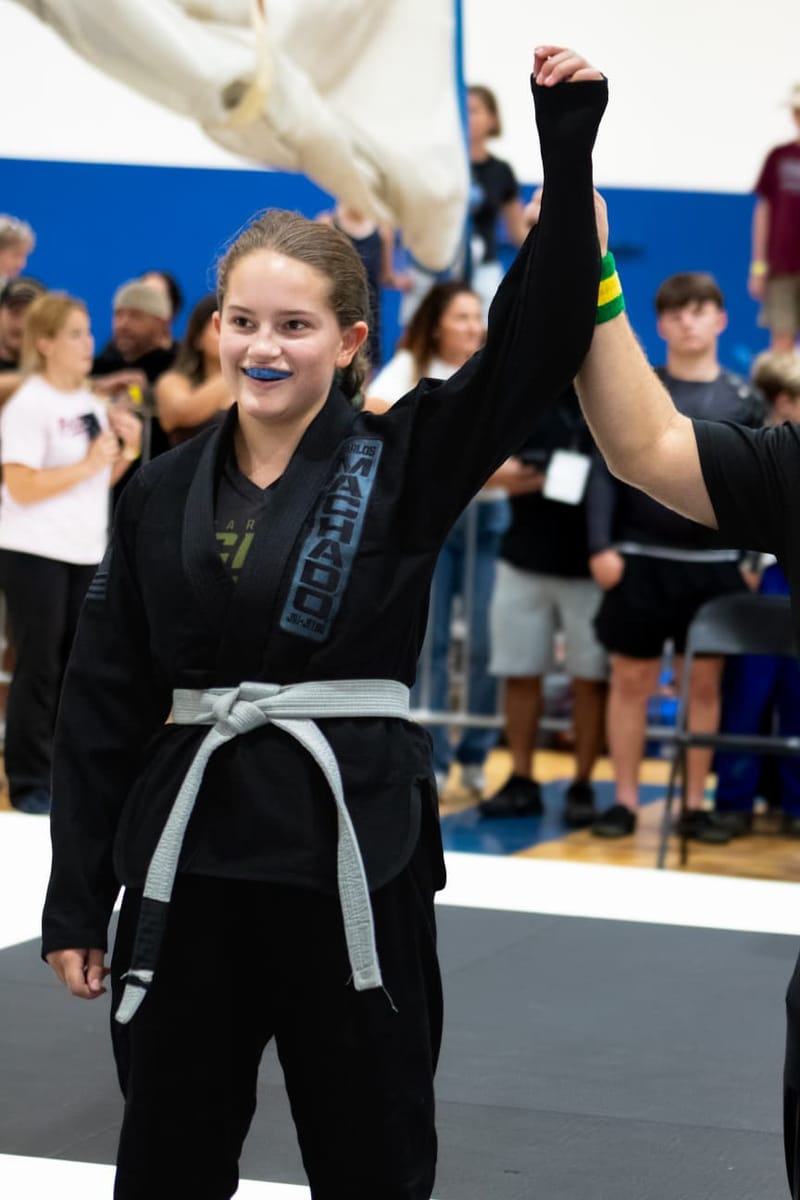 A smiling teenage girl in a black gi has her hand raised in victory after a jiu-jitsu match.