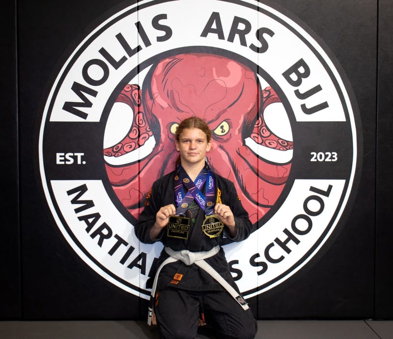 A teen girl in a black gi displays two medals in front of the Mollis Ars BJJ logo wall.