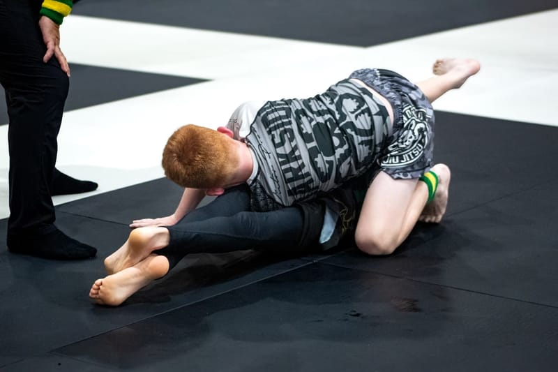 A teenage grappler in grey shorts defends from the turtle position during a no-gi match.