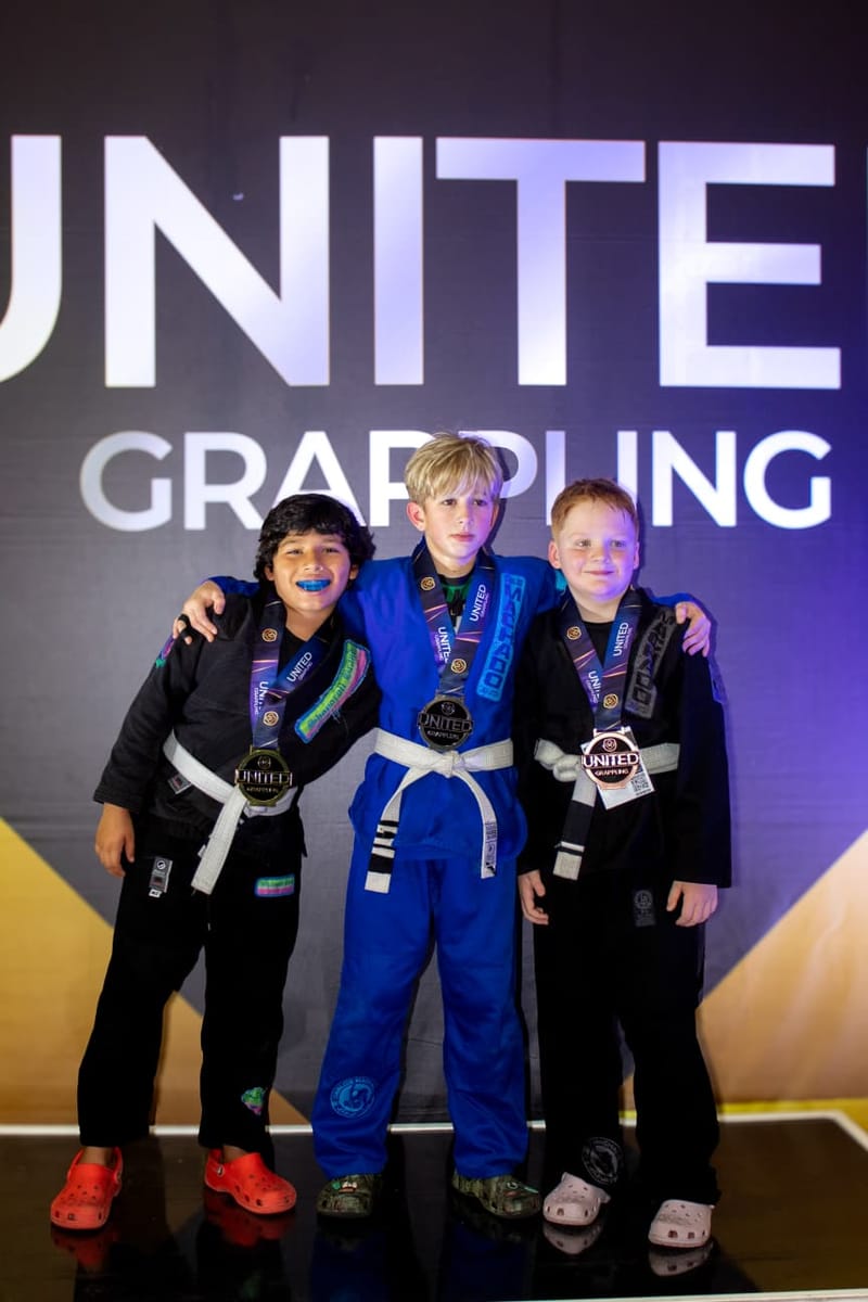 Three young jiu-jitsu students pose with medals in front of a United Grappling event backdrop.