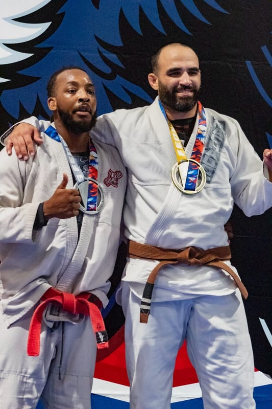 Two adult competitors in white gis pose with their medals in front of the tournament banner.