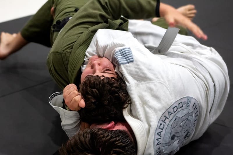 Two adults grapple on the mat as one in a white Machado gi controls from the back.