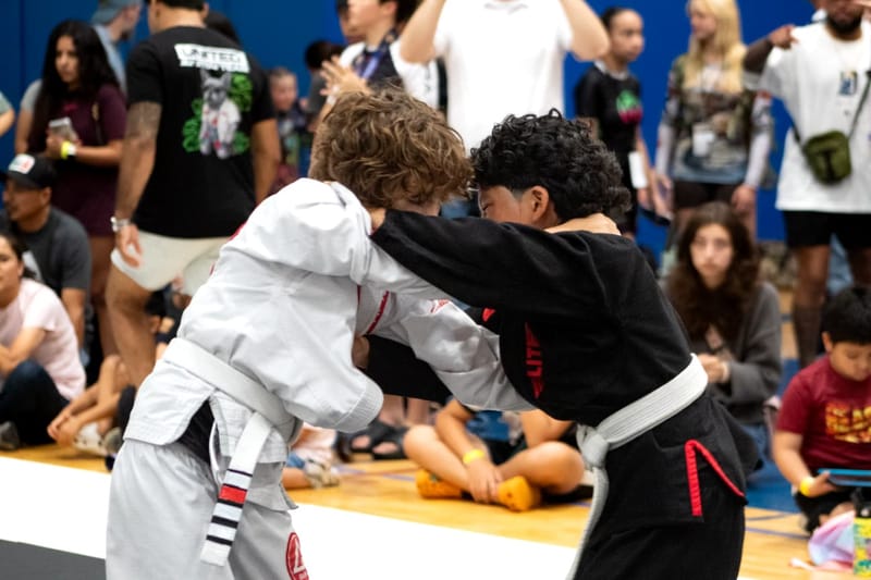 Two boys in white and black gis clinch head-to-head during a jiu-jitsu tournament match.