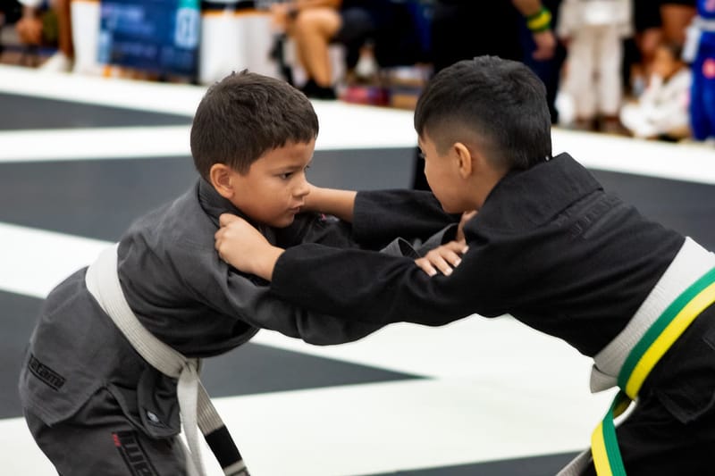 Two young boys grip each other’s collars while facing off in a gi jiu-jitsu match.