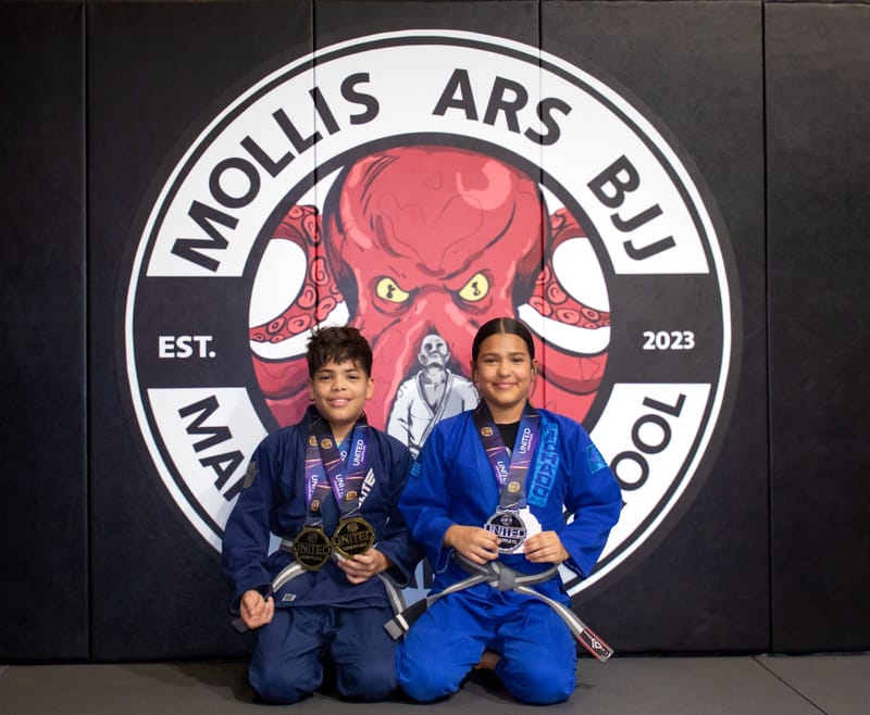 A boy and a girl in blue gis kneel together holding medals at the Mollis Ars BJJ logo wall.