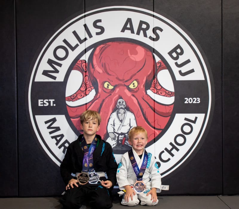 Two young jiu-jitsu students kneel with medals in front of the Mollis Ars BJJ octopus logo wall.