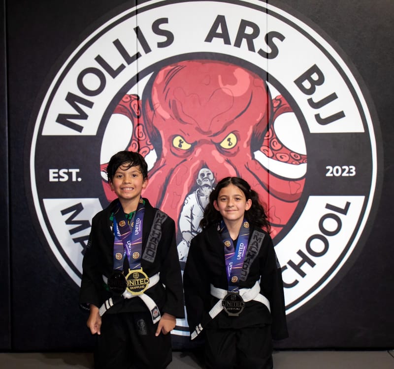 Two young athletes in black gis pose with their medals in front of the Mollis Ars BJJ logo wall.