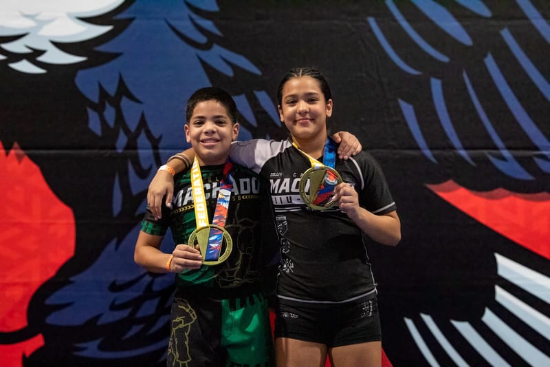 A boy and a girl smile with their medals in front of the tournament banner.