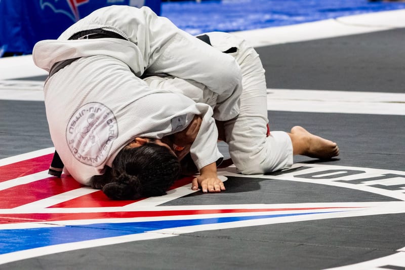 Two women in white gis battle on the ground during a gi match.