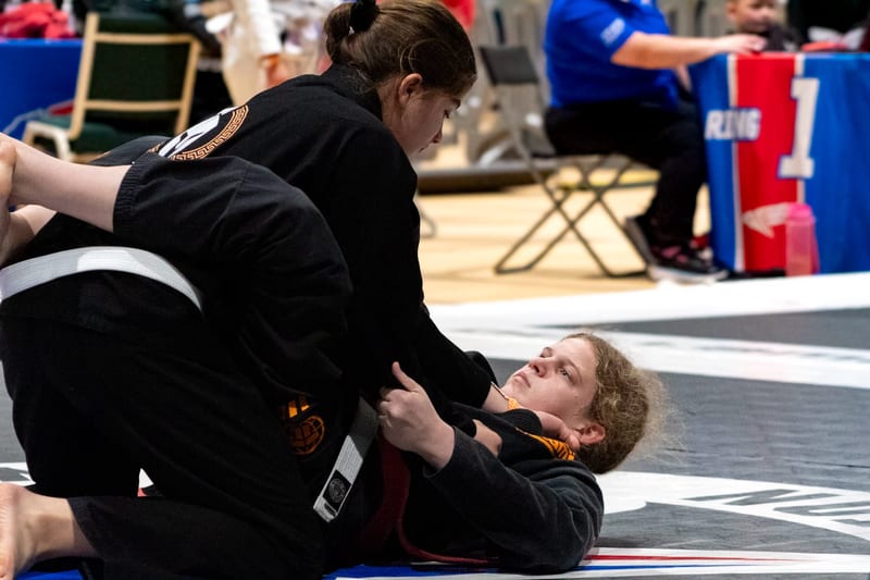A woman in a black gi works to pass her opponent's guard during a women's gi match.