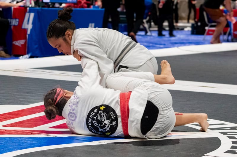 A woman in a white gi holds top position over her opponent during a gi match.