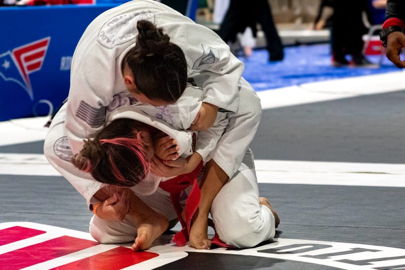 A woman in a white gi attacks from the top as her opponent turtles during a gi match.