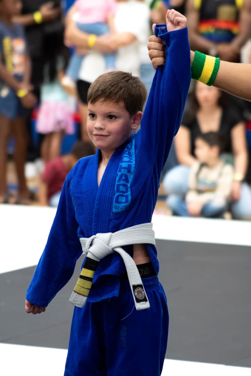 A young boy in a blue gi has his arm raised in victory by a referee at a jiu-jitsu tournament.
