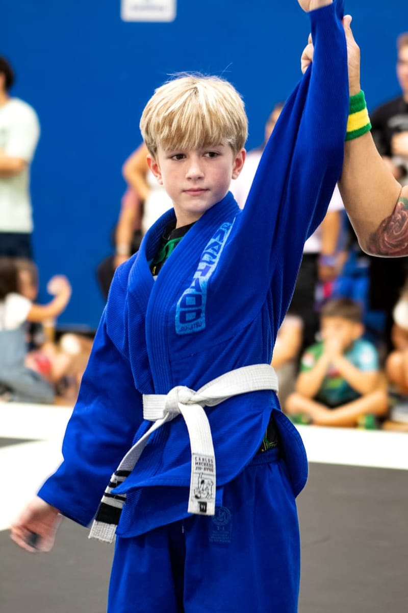 A young boy in a blue gi has his hand raised in victory after his jiu-jitsu match.