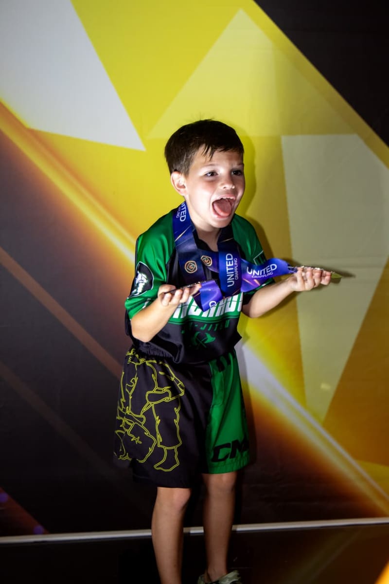 An excited young boy in a green rashguard shows off the medals hanging around his neck.