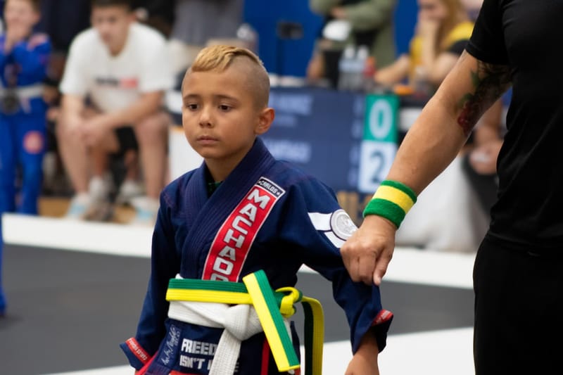 A young boy in a navy Machado gi is led onto the competition mat by his coach.