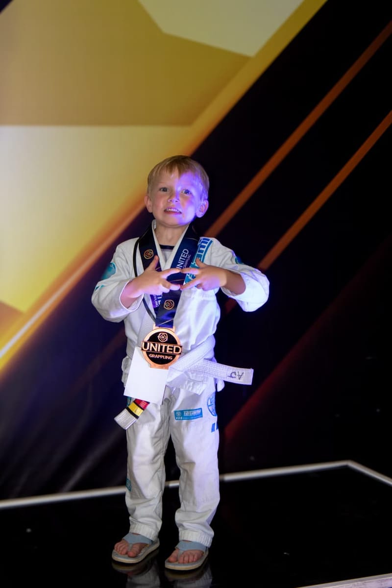 A young boy in a white gi smiles while wearing his United Grappling bronze medal.