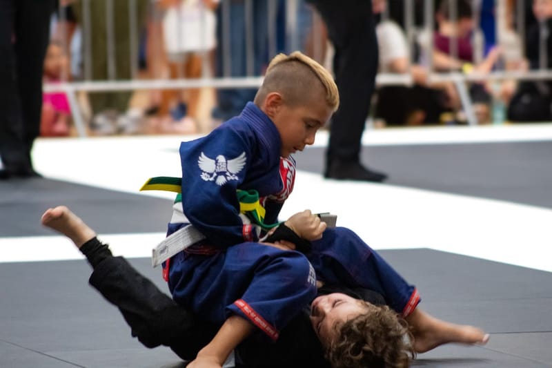 A young boy in a blue gi works to pass the guard of another child during a kids jiu-jitsu match.