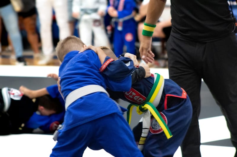 Two young boys in blue gis clinch and fight for a takedown as a referee moves alongside them.