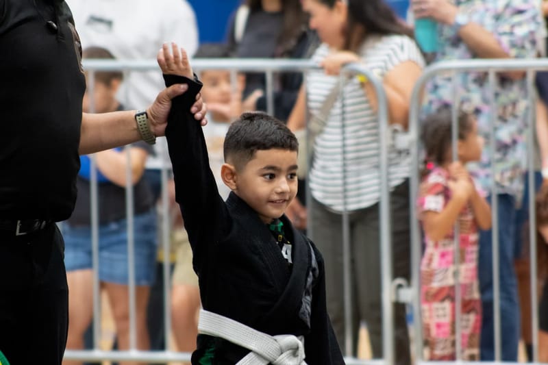 A young boy in a black gi has his hand raised in victory by a coach at a jiu-jitsu tournament.