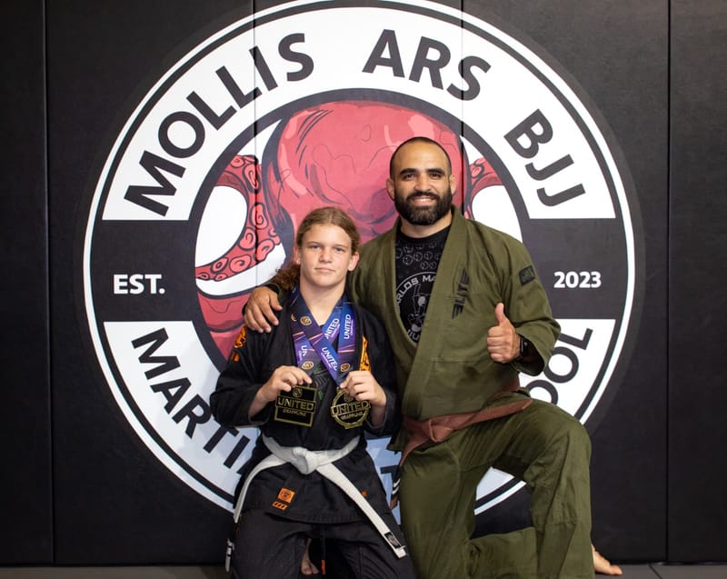 A young competitor in a black gi holds two gold medals next to a smiling brown-belt coach at the logo wall.