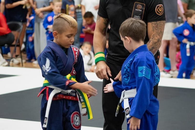 Two young boys adjust their belts beside a referee before a kids jiu-jitsu match.