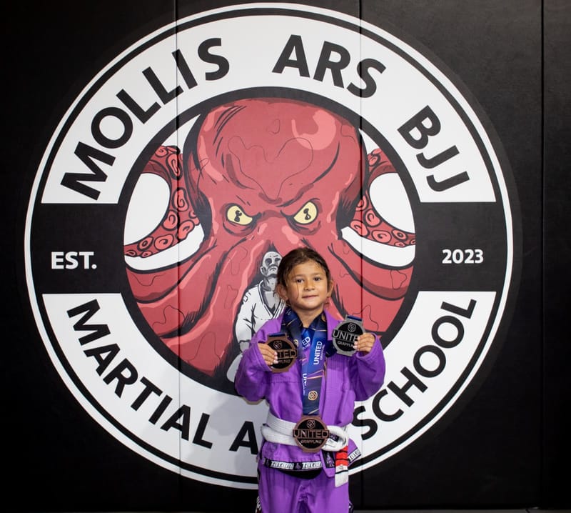 A small girl in a purple gi proudly holds up three medals at the Mollis Ars BJJ logo wall.