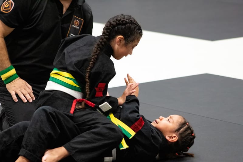 A young girl with braided hair controls her opponent from the top during a girls jiu-jitsu match.