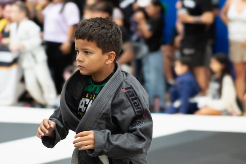 A focused young boy in a gray gi walks across the mat with spectators in the background.