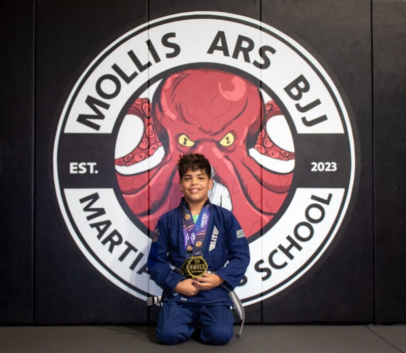 A smiling boy in a blue gi holds his gold medal in front of the Mollis Ars BJJ logo wall.