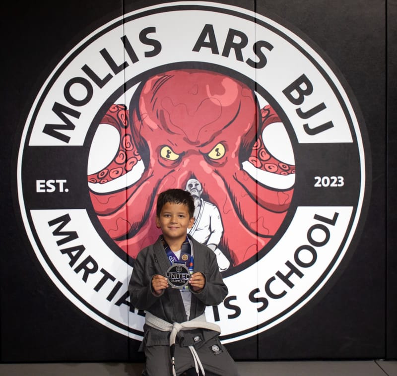 A smiling boy in a grey gi holds up his medal in front of the Mollis Ars BJJ logo wall.