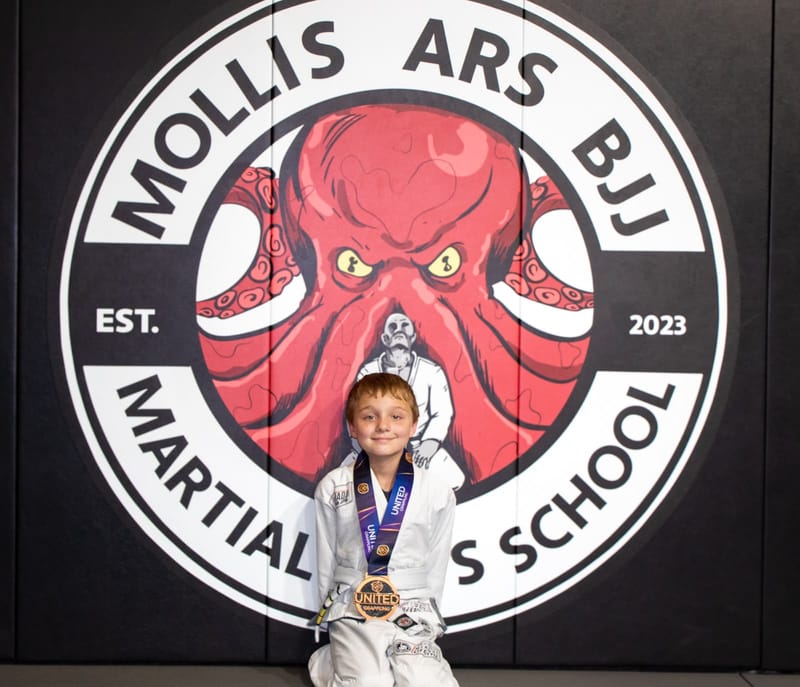A smiling boy in a white gi holds his medal in front of the Mollis Ars BJJ logo wall.