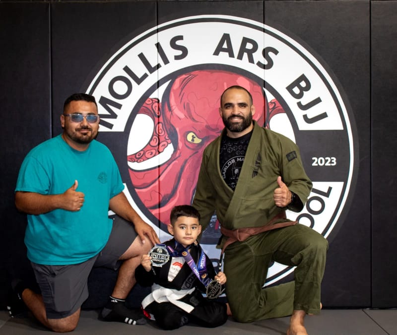A young boy in a black gi holds awards while his father and a brown-belt coach kneel beside him.