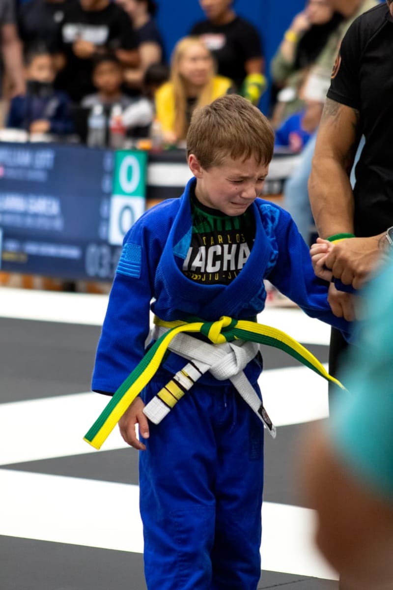 A boy in a blue gi tears up beside a referee after a hard-fought jiu-jitsu match.